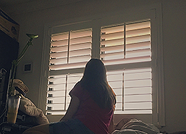 A picture of Estefi in a darkened room, silhouetted against a window with the wooden blinds turned open letting in some light.