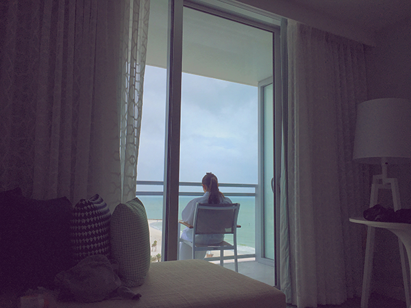 Dimly lit photo, with Estefi set at the center, head bowed, framed by a blacony door, with the ocean in view in the distance.